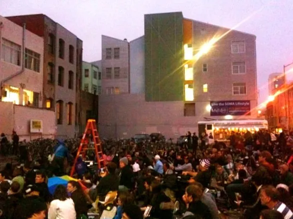 Crowd sitting on the ground watching a movie with SOMA Residences in San Francisco in the background.
