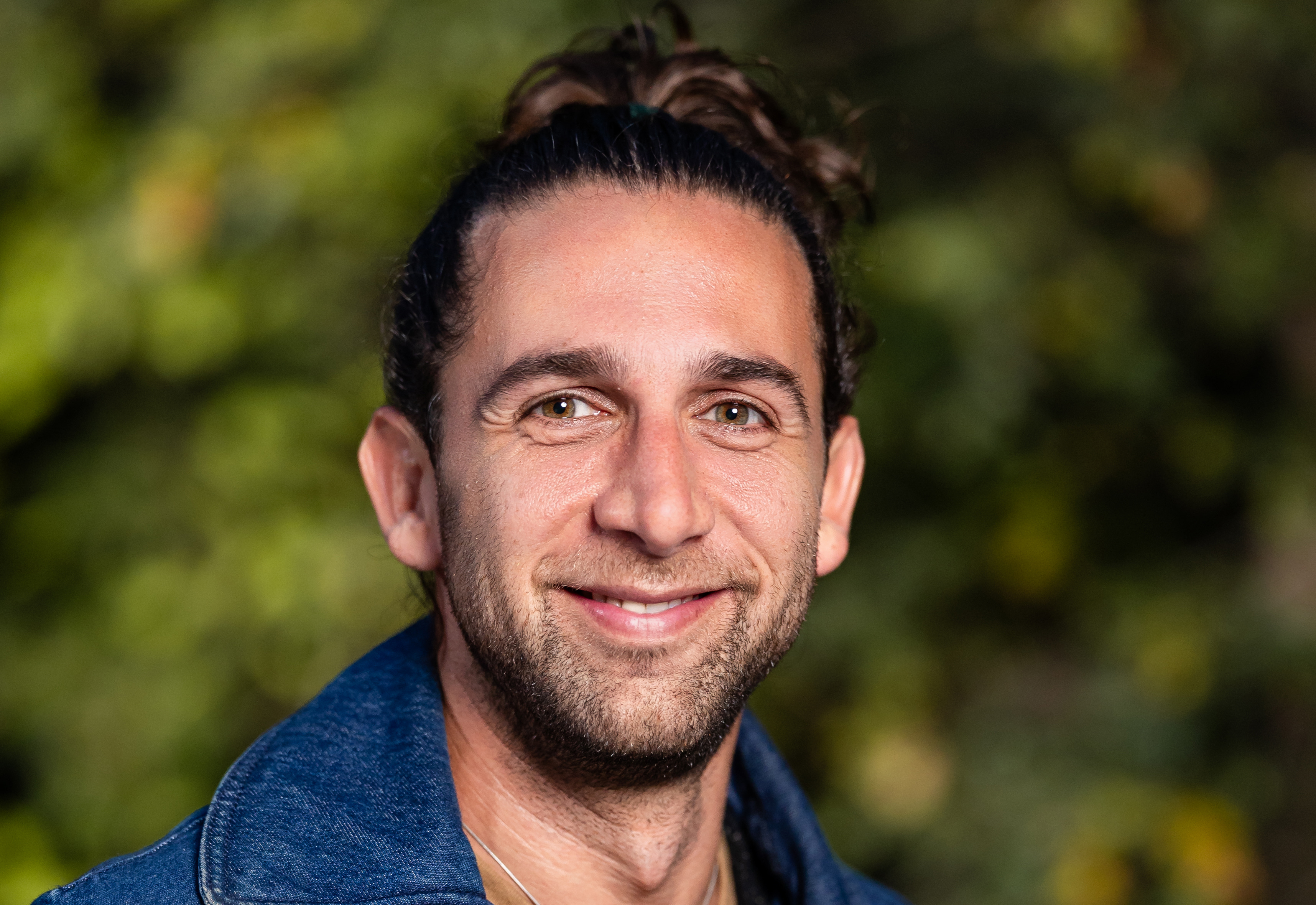 Headshot of man with brown hair in bun wearing denim jacket