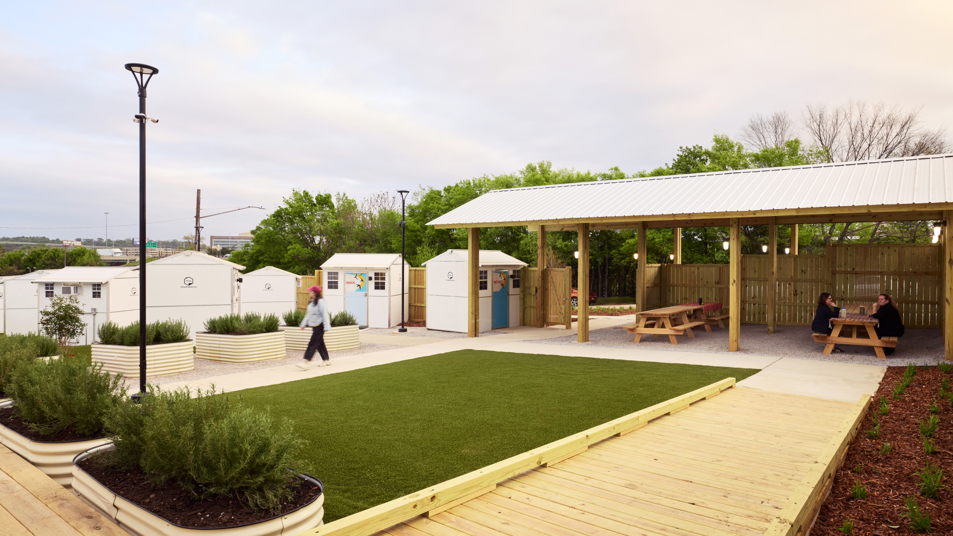 Raised wooden pathways surround planters and a patch of grass, with small structures in the background