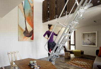 Interior view of the living room and a woman climbing up metal stairs to the third level inside Zero Cottage in San Francisco.