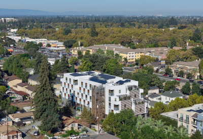 Aerial view of front corner of white apartment building in cityscape with hills and blue sky in background