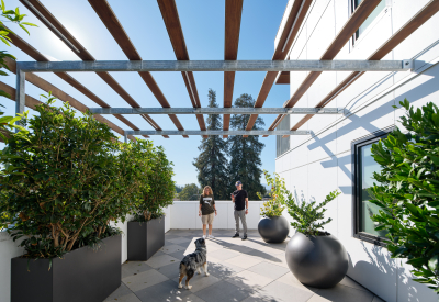 Two people and a dog on a patio with planters and a wood trellis overhead
