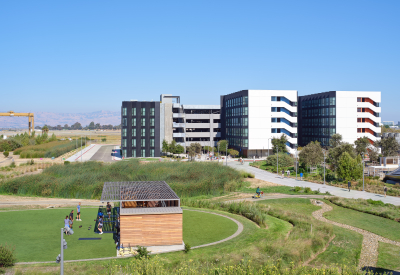 Large gray and white building with small building with wood siding in a park in foreground against blue sky