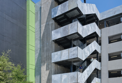 Gray building with stair and green vertical band against a blue sky with cars on road in front