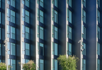 Gray sawtooth building with trees and paved park in foreground