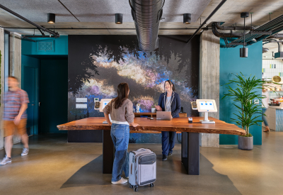 Two people facing each other across a wood slab reception desk with teal and black decorative wall in background 