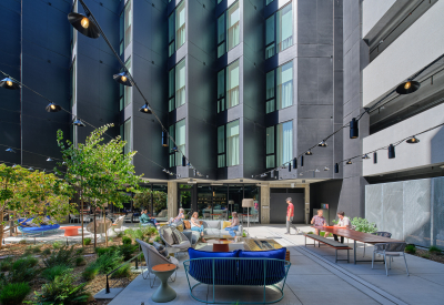 Gray sawtooth building in background with people gathered in exterior courtyard with overhead lighting