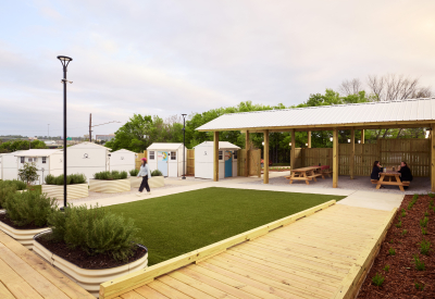 Raised wooden pathways surround planters and a patch of grass, with small structures in the background