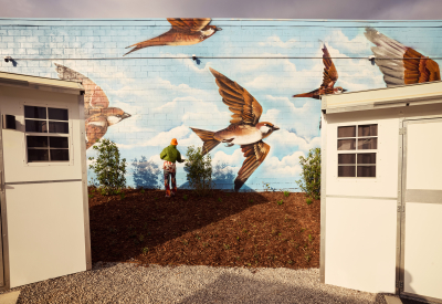 An artist paints a mural of oversize sparrows on a pale blue sky on a wall behind two small structures