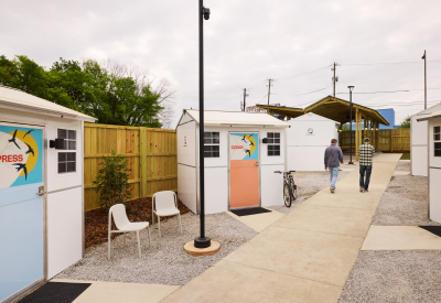 Two people walk down a path past small structures with seating out front