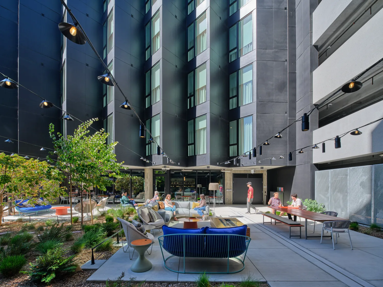 Gray sawtooth building in background with people gathered in exterior courtyard with overhead lighting