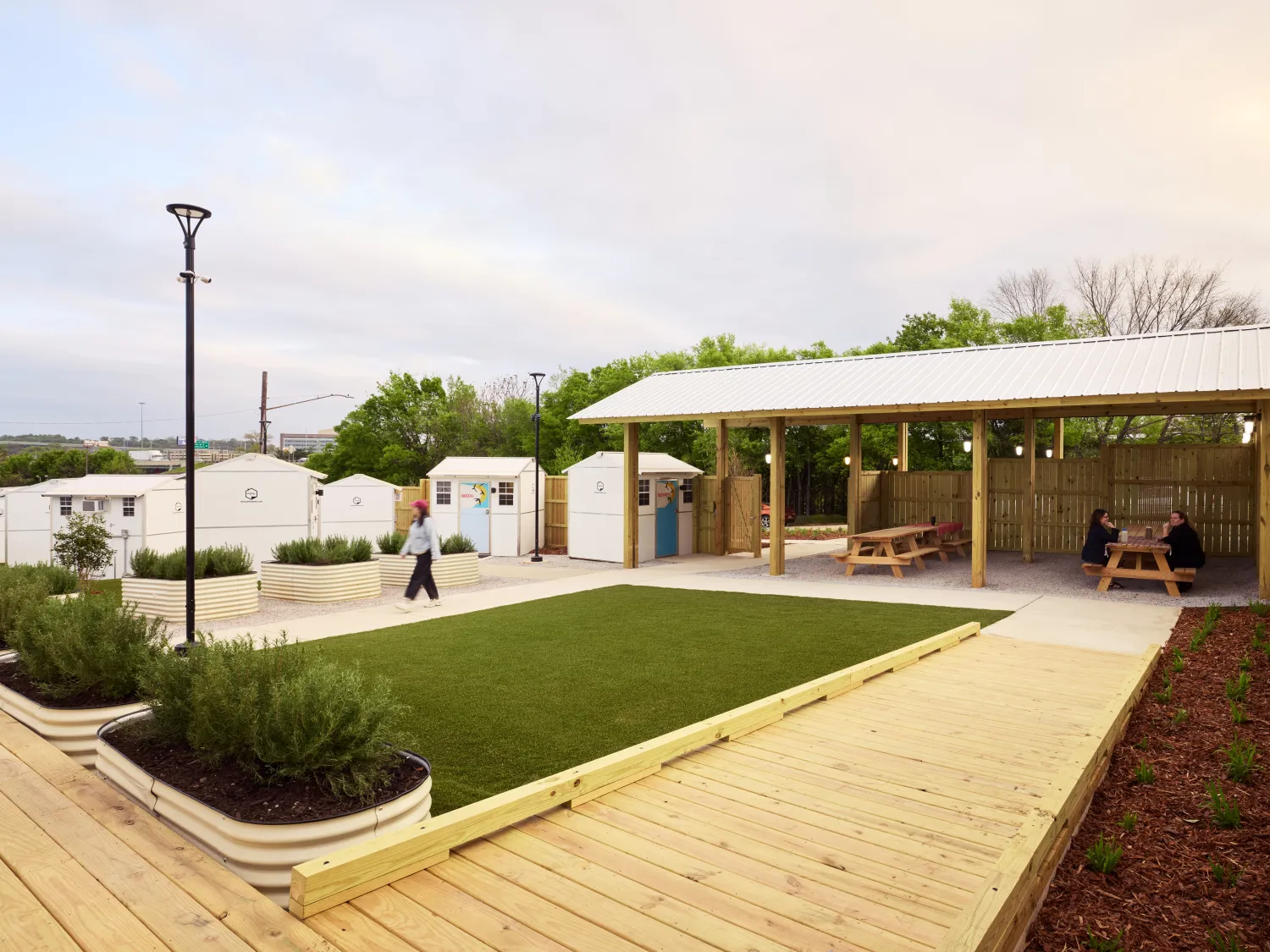 Raised wooden pathways surround planters and a patch of grass, with small structures in the background