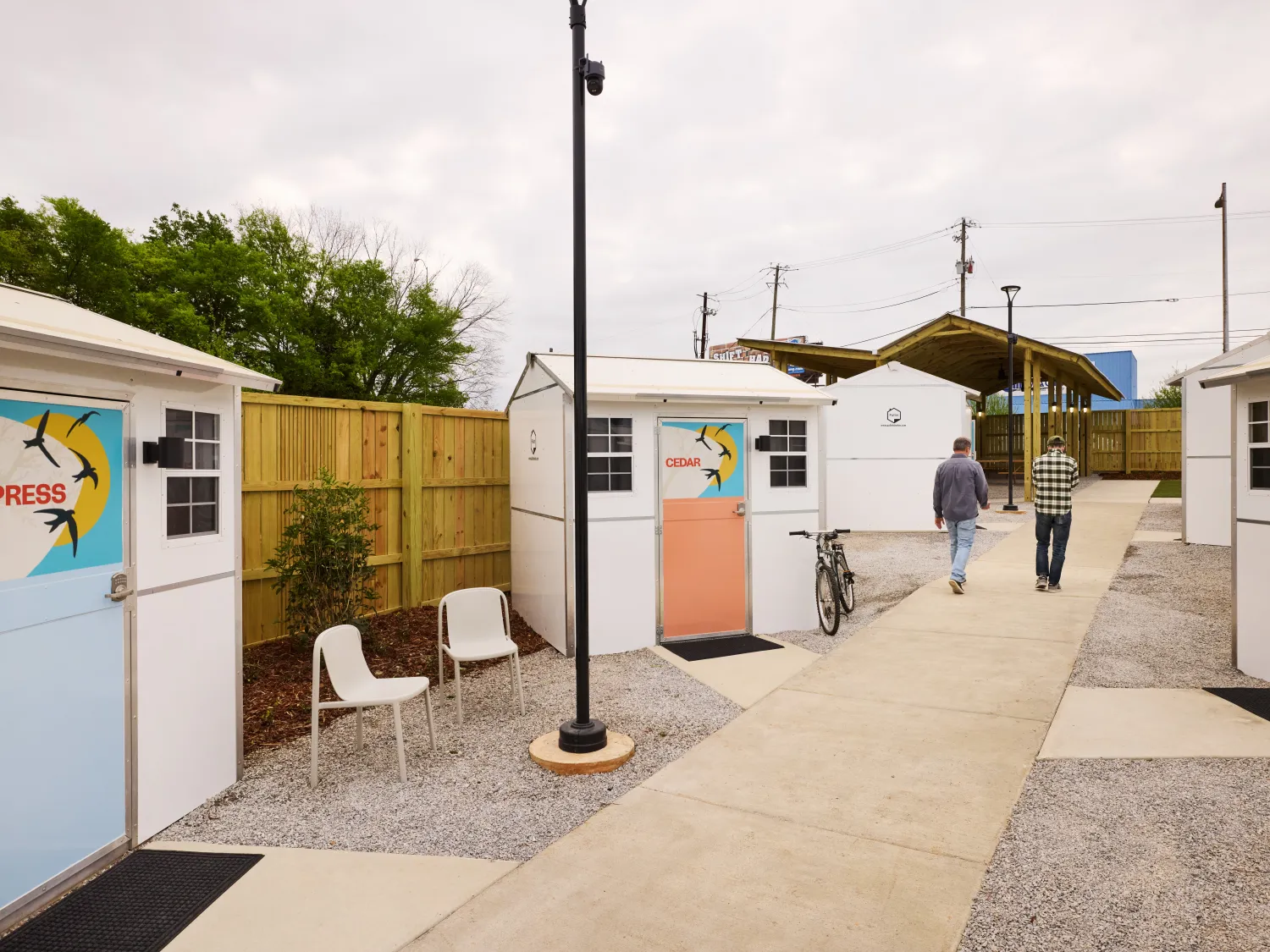 Two people walk down a path past small structures with seating out front