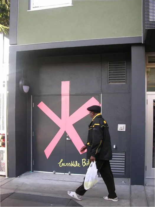 Man waling past a painted mural on the dark facade of Curran House showing a big pink asterisk with a handwritten legend in yellow reading "Incredible but true"