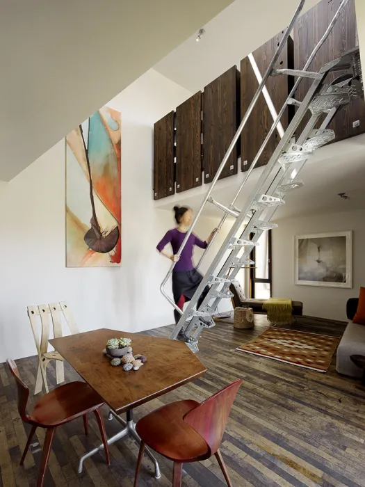 Interior view of the living room and a woman climbing up metal stairs to the third level inside Zero Cottage in San Francisco.