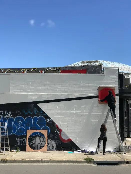 Two people on a ladder outside of David Baker Architects office to put up art on the wall.