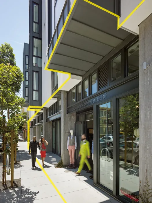 View of sidewalk at mixed-use urban building showing property line marked in yellow, with the building pulled back from the property line and active uses at edge.