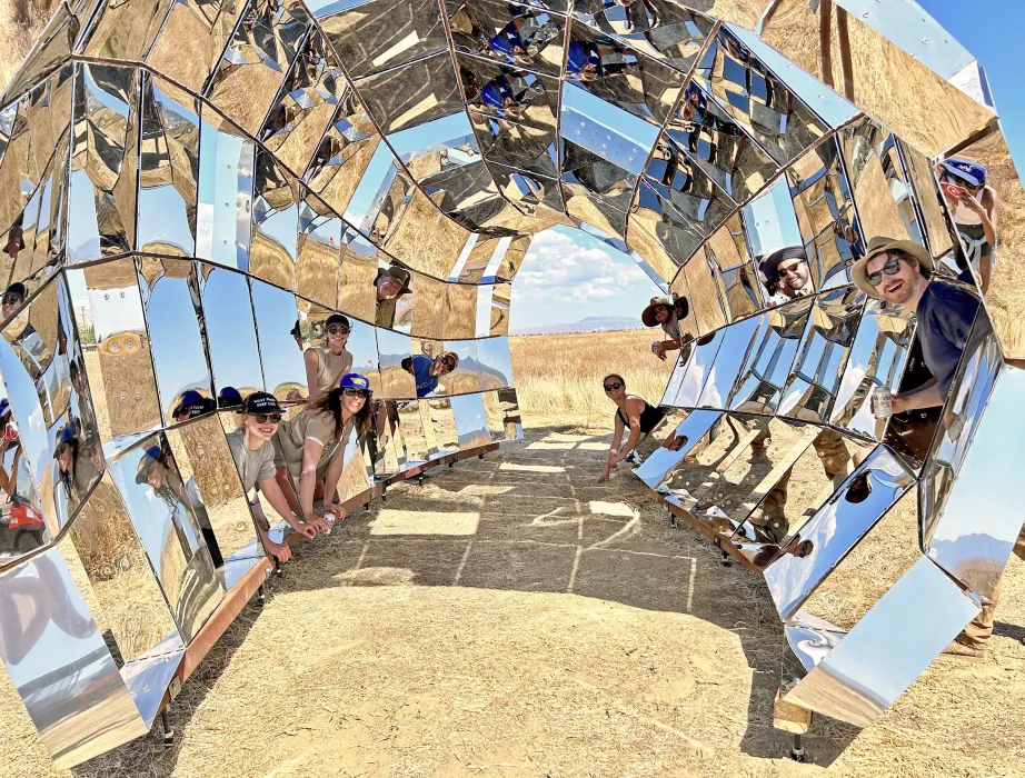 A group of people, the DBA_Lab team poking their heads inside peepSHOW, in the desert in New Cuyama, California.