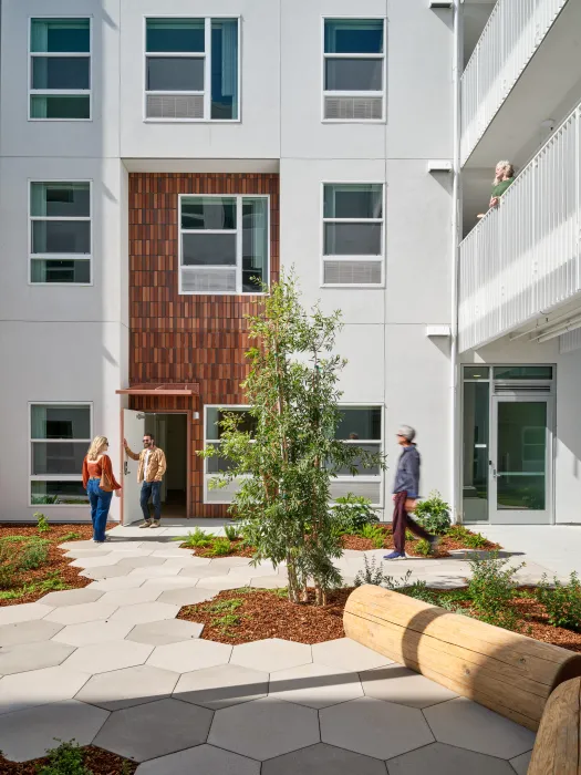 Apartment entry with two people talking viewed from a courtyard with hexagonal concrete tiles and log benches