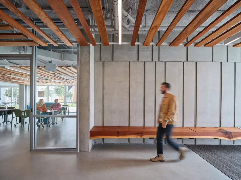 Man walking through entry hallway with wood ceiling and bench, textured concrete walls, and view into community room