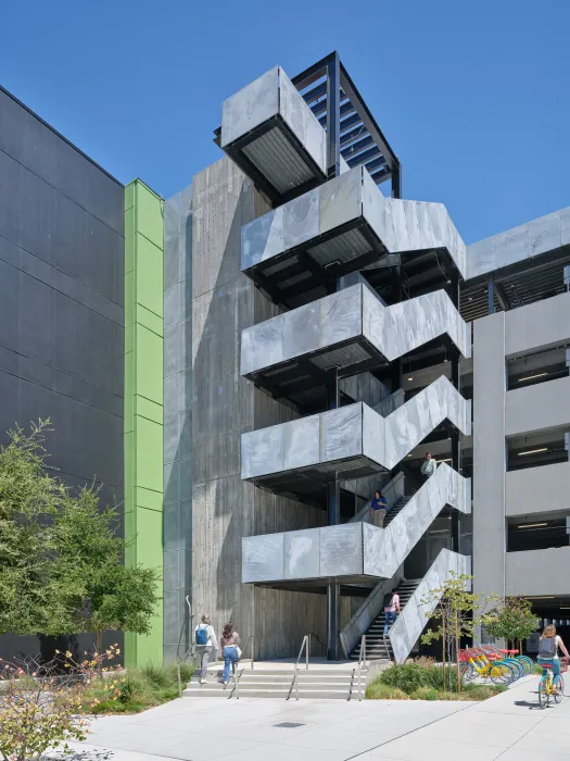 Gray building with stair and green vertical band against a blue sky with cars on road in front
