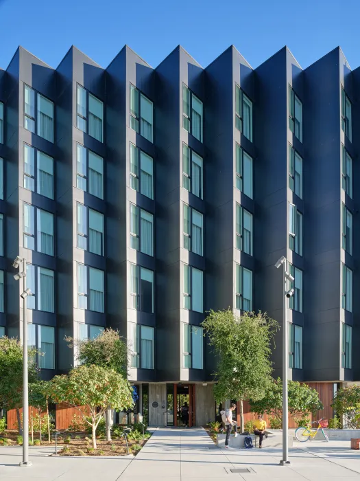Gray sawtooth building with trees and paved park in foreground
