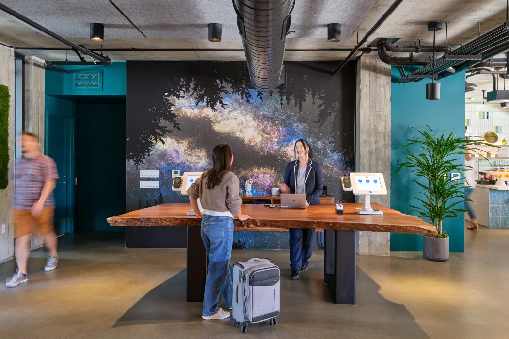 Two people facing each other across a wood slab reception desk with teal and black decorative wall in background 