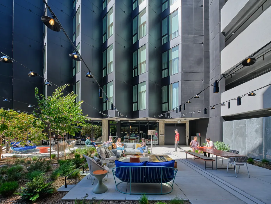 Gray sawtooth building in background with people gathered in exterior courtyard with overhead lighting