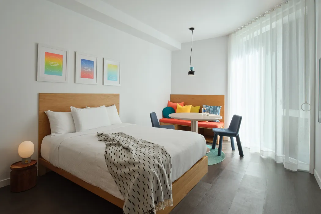 White bedroom with wooden built in seating and round table in background and three framed prints above a wood bed