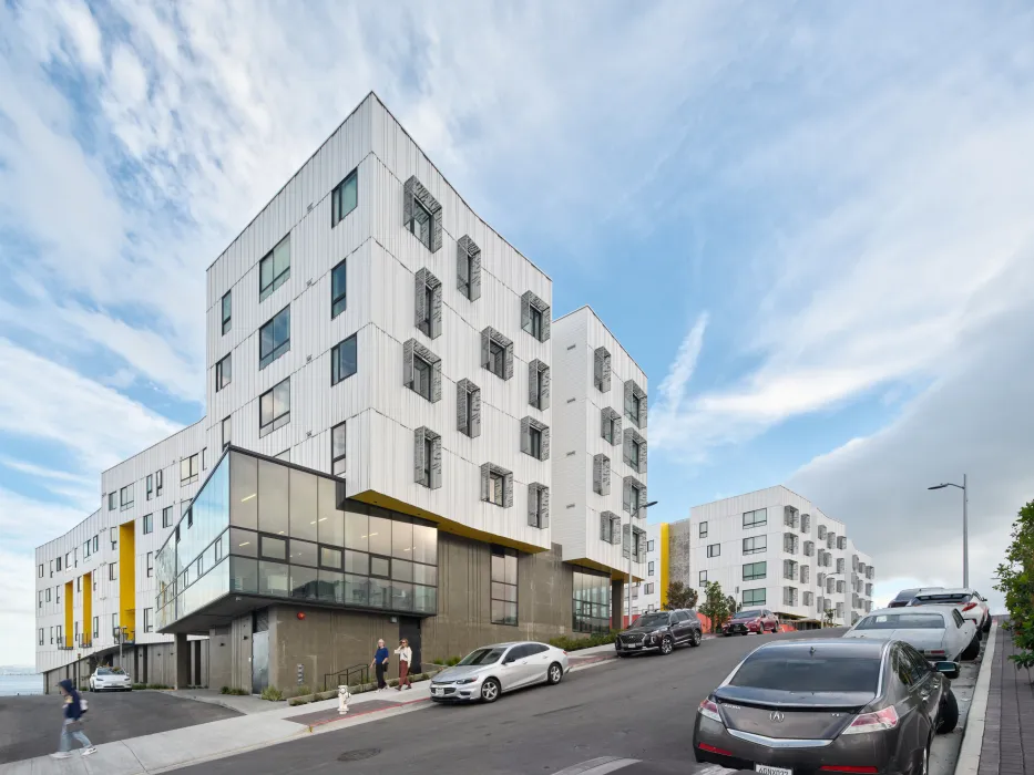 Street view of white apartment building with cars and pedestrians