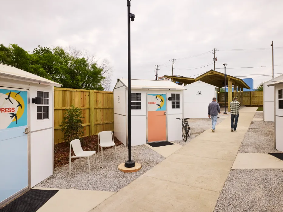 Two people walk down a path past small structures with seating out front